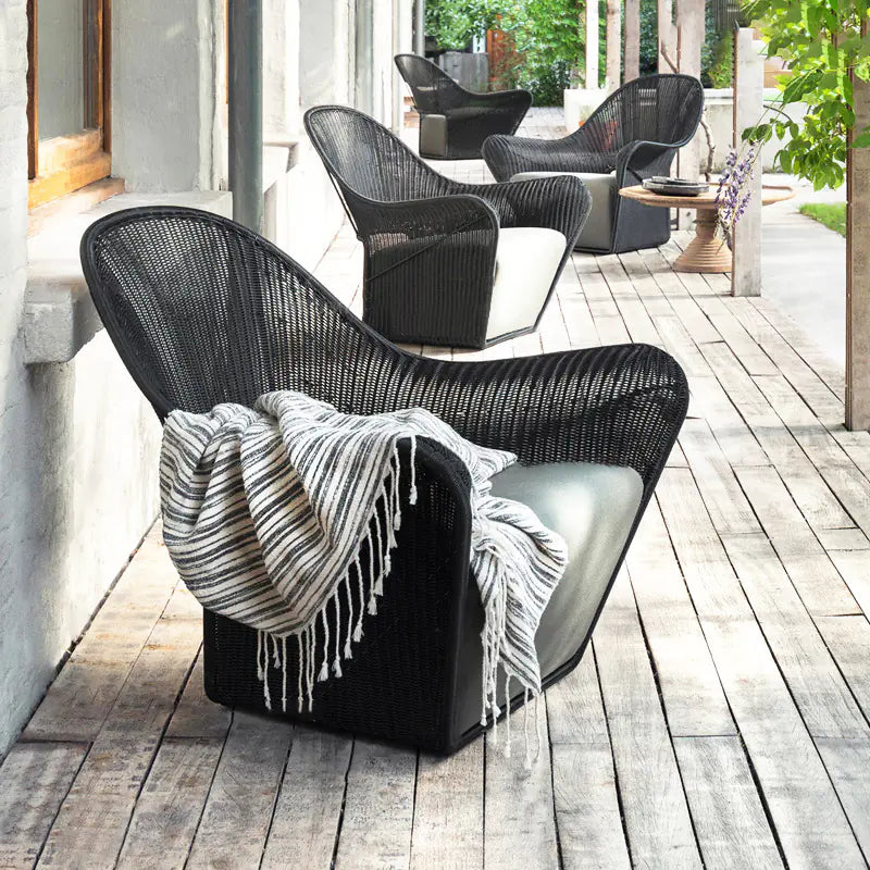 Outdoor single rattan sofa in black color placed on a wooden deck with a striped beige and black throw over the backrest.